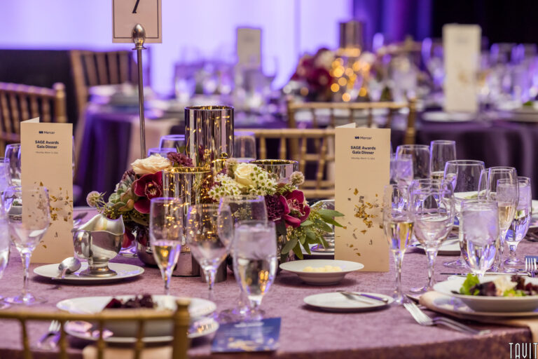 A formal banquet table is elegantly set for a corporate event, featuring glassware, plates, menus, candles, and a floral centerpiece, surrounded by gold chairs and softly lit with purple ambient lighting.