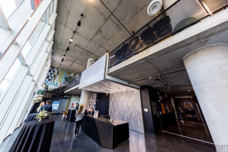 A modern, industrial-style lobby with high ceilings, concrete surfaces, large windows, and a geometric reception desk—an ideal setting for a corporate event. People gather near the desk while colorful wall art decorates the space.