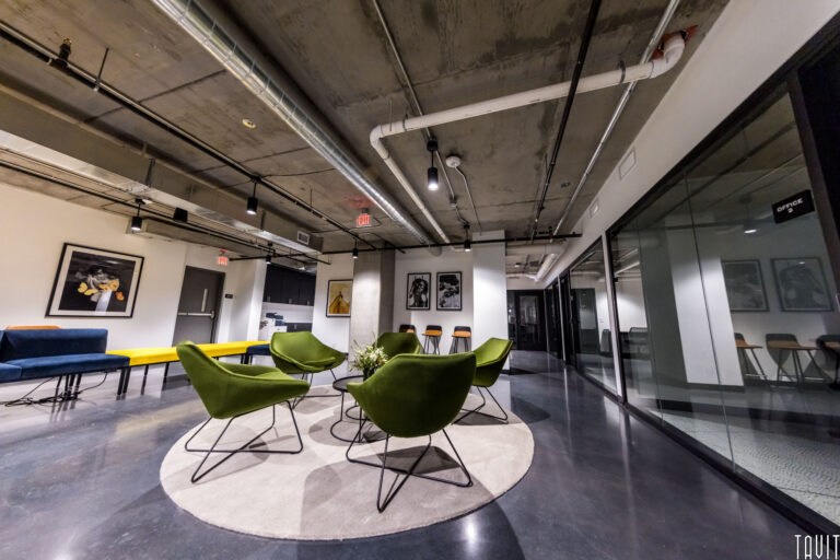 Modern office lounge ideal for a seminar or corporate event, featuring four green chairs around a round rug, yellow bench, artwork on white walls, glass-walled meeting rooms, and an exposed concrete ceiling with visible pipes.