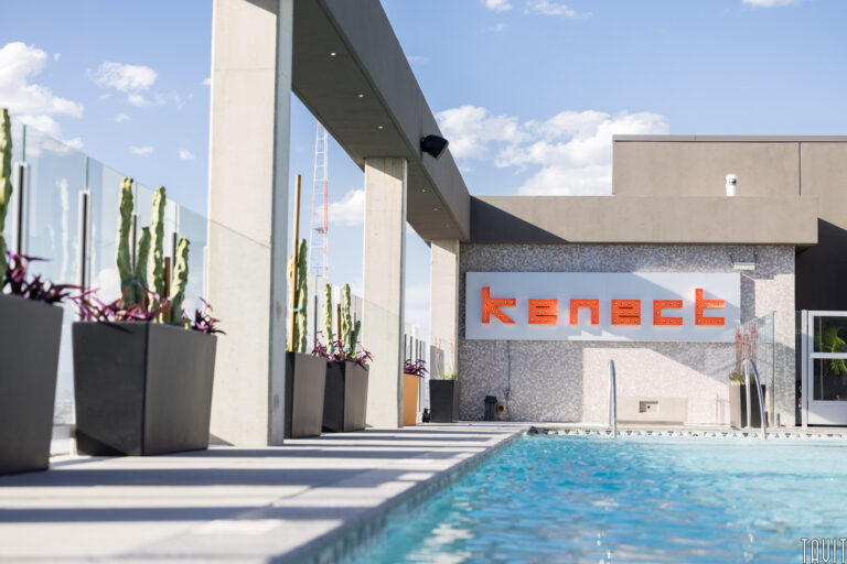 A modern rooftop pool with potted cacti along one side and a wall featuring the orange "kenect" sign—an ideal spot for a seminar or corporate event under a clear sky with a few clouds.