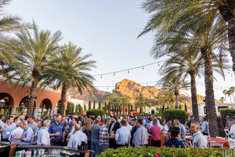 A large group of people gather outdoors at a corporate event among palm trees, with string lights overhead and mountains in the background at sunset. Tables with drinks and food are visible in the foreground.