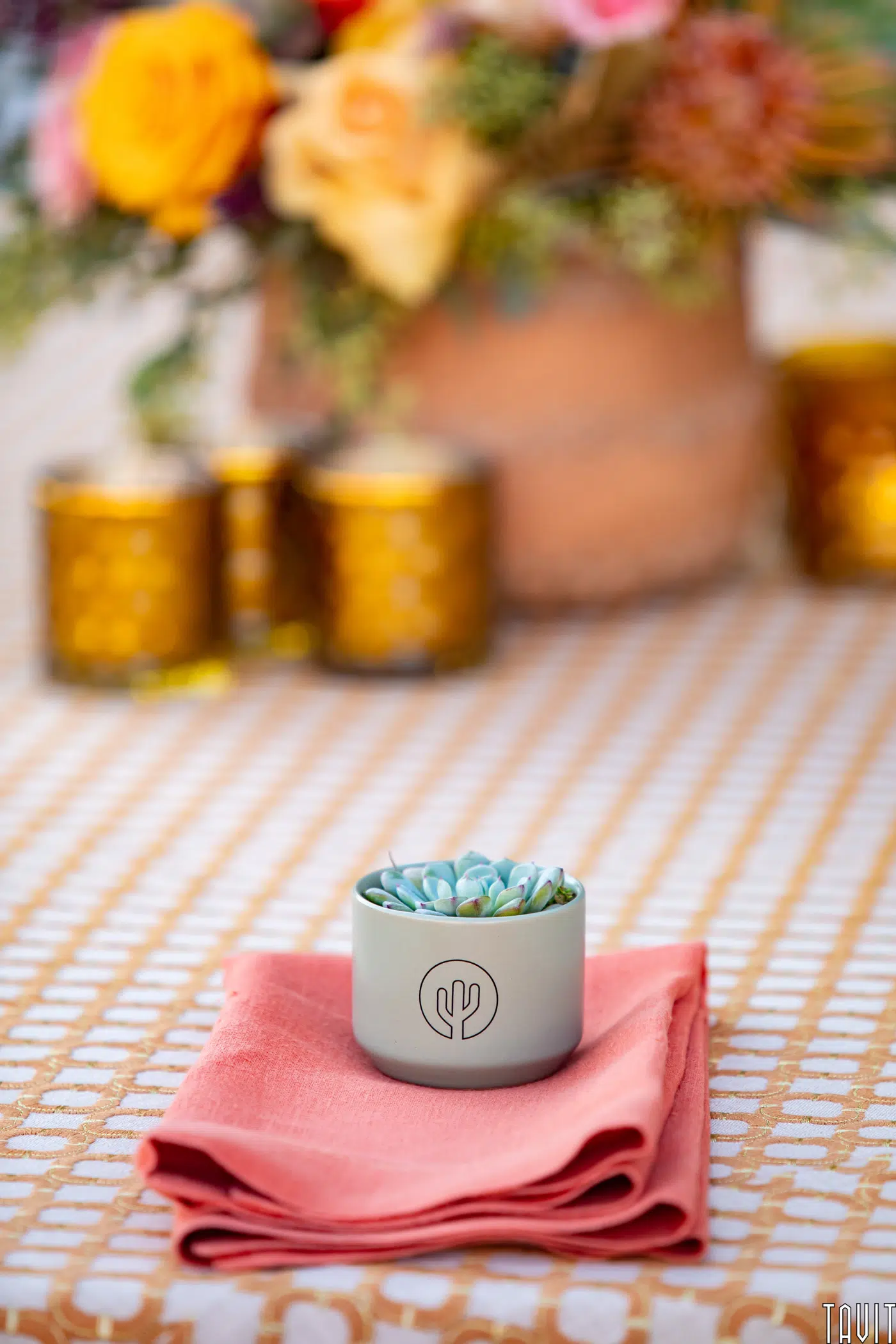 A small succulent in a white pot with a cactus design sits on a folded coral napkin atop a patterned tablecloth, adding charm to your seminar or corporate event. In the background are yellow glass candle holders and a blurred vase of colorful flowers.