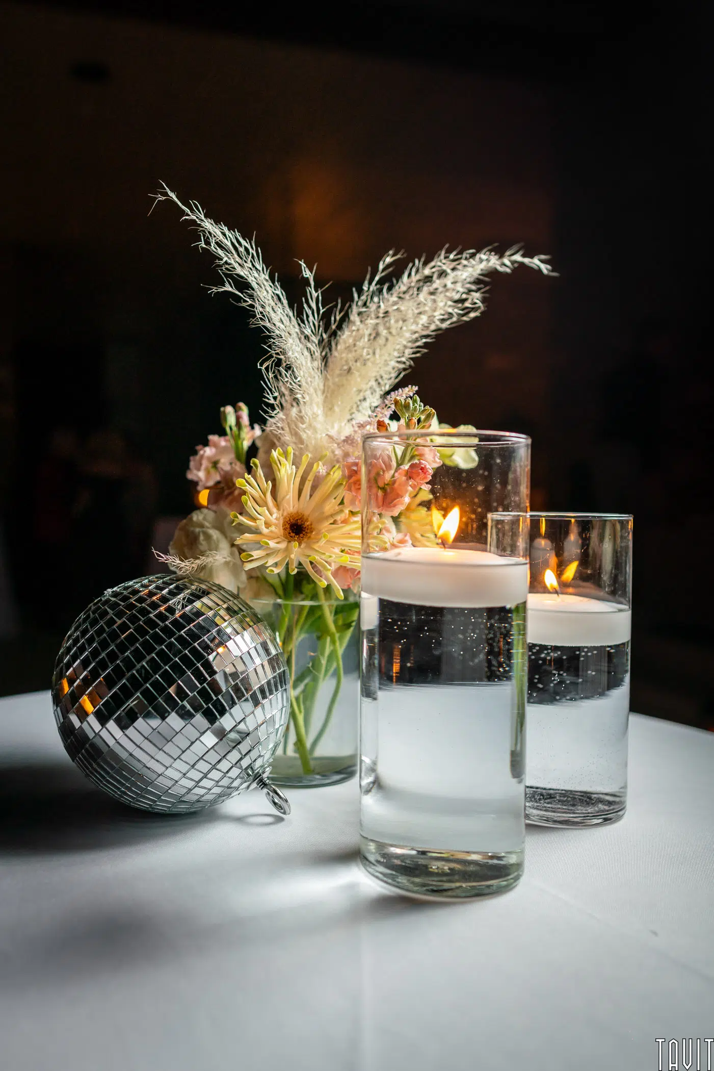A table centerpiece perfect for a corporate event, featuring a bouquet of flowers in a jar, two tall glass candles floating in water, and a small disco ball, all set on a crisp white tablecloth.