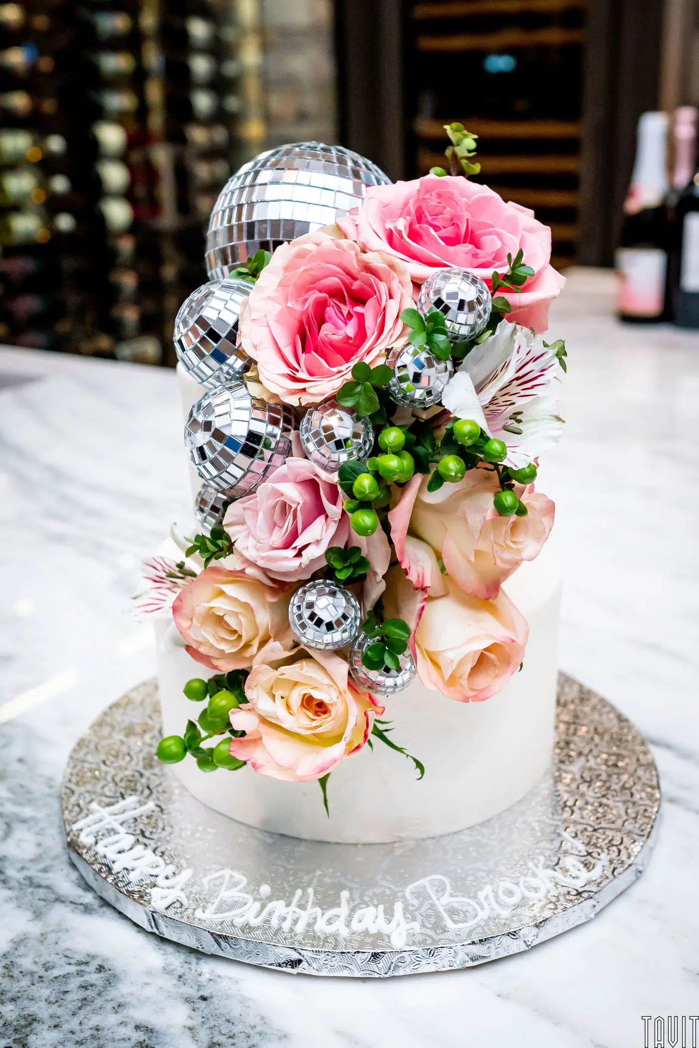 A white birthday cake is decorated with pink and peach roses, greenery, and small silver disco balls cascading down one side—perfect for a special birthday or a sophisticated seminar celebration. "Happy Birthday Brooke" is written on the silver board.