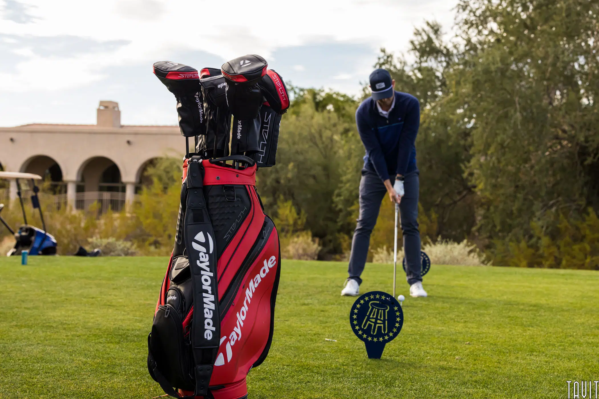 A red TaylorMade golf bag with clubs stands on the grass at a corporate event while a golfer in blue prepares to swing in the background, surrounded by trees and a building under a partly cloudy sky.