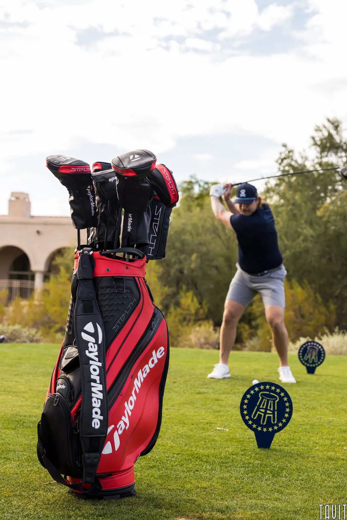 A red and black TaylorMade golf bag is in focus on a grassy course during a corporate event, while a golfer in a blue shirt and gray shorts swings a club in the blurred background. Blue and yellow event markers are on the green.