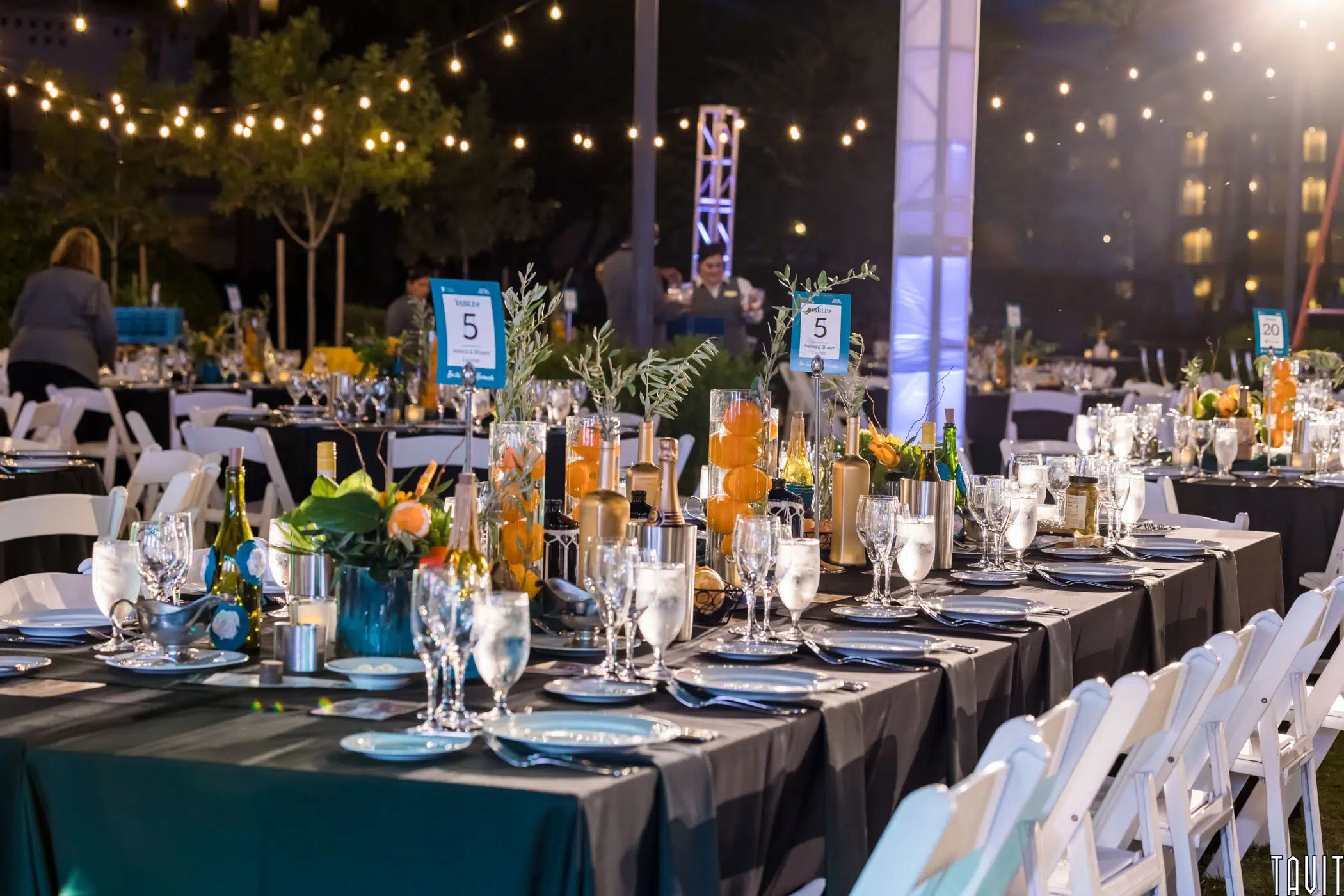 Elegant outdoor corporate event dinner setup at night with string lights overhead, neatly arranged tables with glassware, plates, candles, and floral centerpieces. Numbered table signs are visible as guests mingle during the seminar in the background.