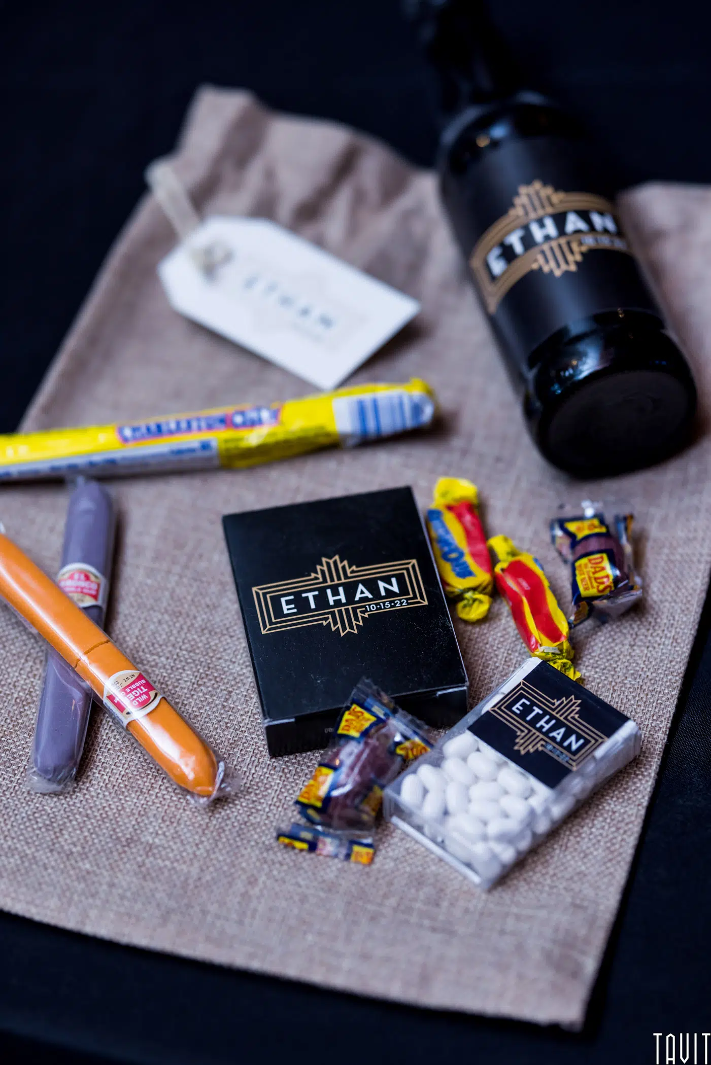 A burlap mat with various items labeled "Ethan"—a black bottle, a box, a white mint box, a name tag, two cigar-shaped candies, and assorted sweets—neatly arranged as ideal giveaways for a corporate event or seminar.