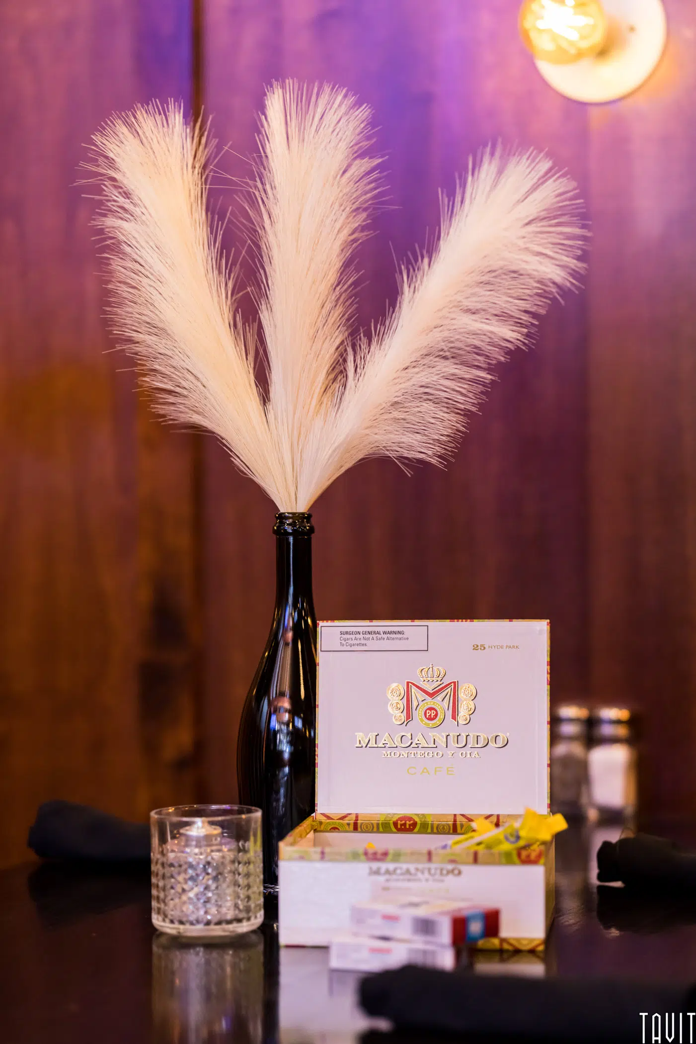 A dark bottle holding pampas grass sits on a table next to a candle, a box of Macanudo cigars, and salt and pepper shakers—perfect décor for an intimate seminar or stylish corporate event—against a wooden wall with warm light above.