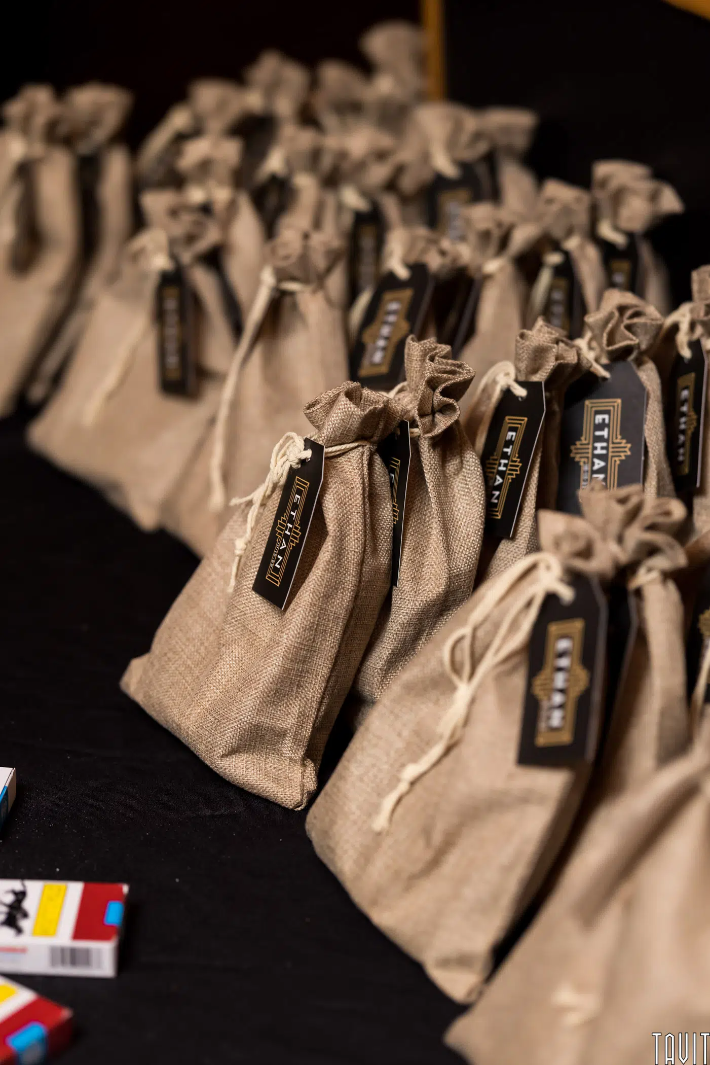 A group of small burlap gift bags with drawstrings and black tags, perfect for a seminar or corporate event, are arranged in rows on a black surface. Some colorful rectangular boxes are partially visible in the lower left corner.