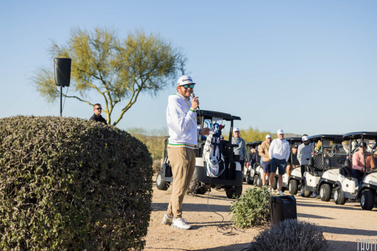 A man in sunglasses and a white jacket speaks into a microphone outdoors at a corporate event, standing near golf carts and a speaker, with several people and a tree visible in the background on a sunny day.