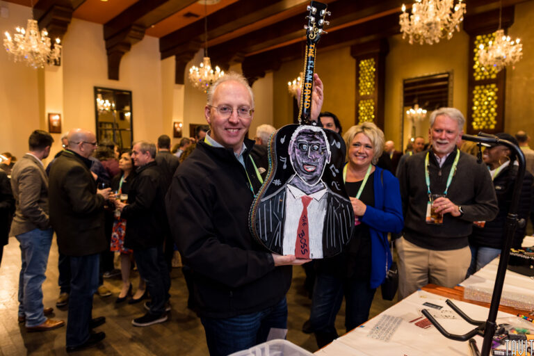 A smiling man holds a painted guitar with a face and suit design at an indoor corporate event, surrounded by people mingling in a warmly lit, elegant room with chandeliers.