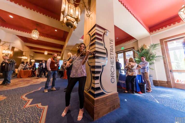 A woman playing the violin stands by a decorated pillar at a bustling corporate event in a large, ornate room, with people mingling and booths in the background. The pillar features “ORCHESTRATE” and stylized music-themed graphics.