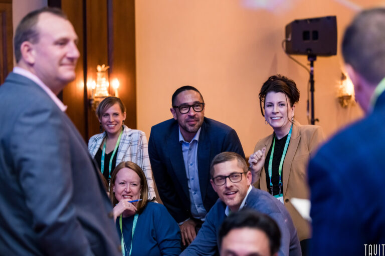 A group of six adults, dressed in business attire and wearing lanyards, smile and engage with someone off-camera in a warmly lit conference room during a corporate event.