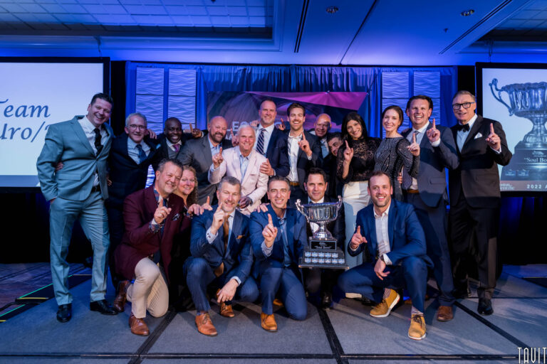 A group of people in formal attire smile and pose together on stage at a corporate event, holding up their index fingers. They stand around a large trophy, celebrating a team achievement at this indoor celebration.