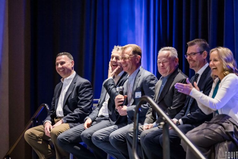 Six professionally dressed people sit on a stage under blue lighting, smiling and laughing at a corporate event. One man holds a microphone while a woman on the end claps enthusiastically.