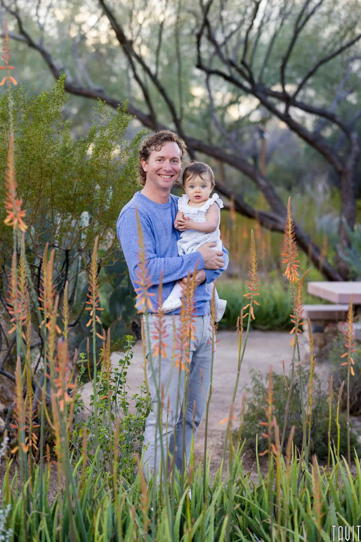 Man smiling holding baby behind desert plants