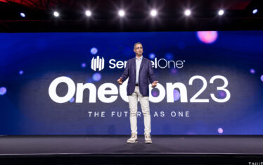 presenter standing on stage in front of SettinelOne's OneCon23 slide