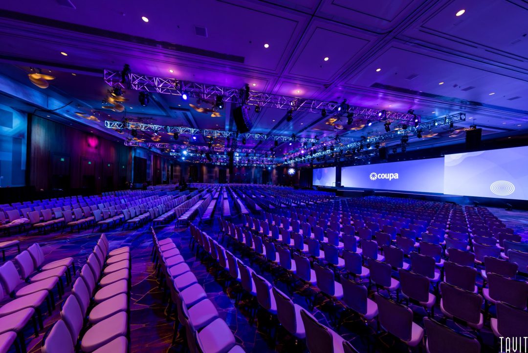 Rows of empty chairs face a large stage with multiple screens displaying the Coupa logo, under vibrant purple and blue lighting, setting the scene for a corporate event or seminar in a spacious conference hall.