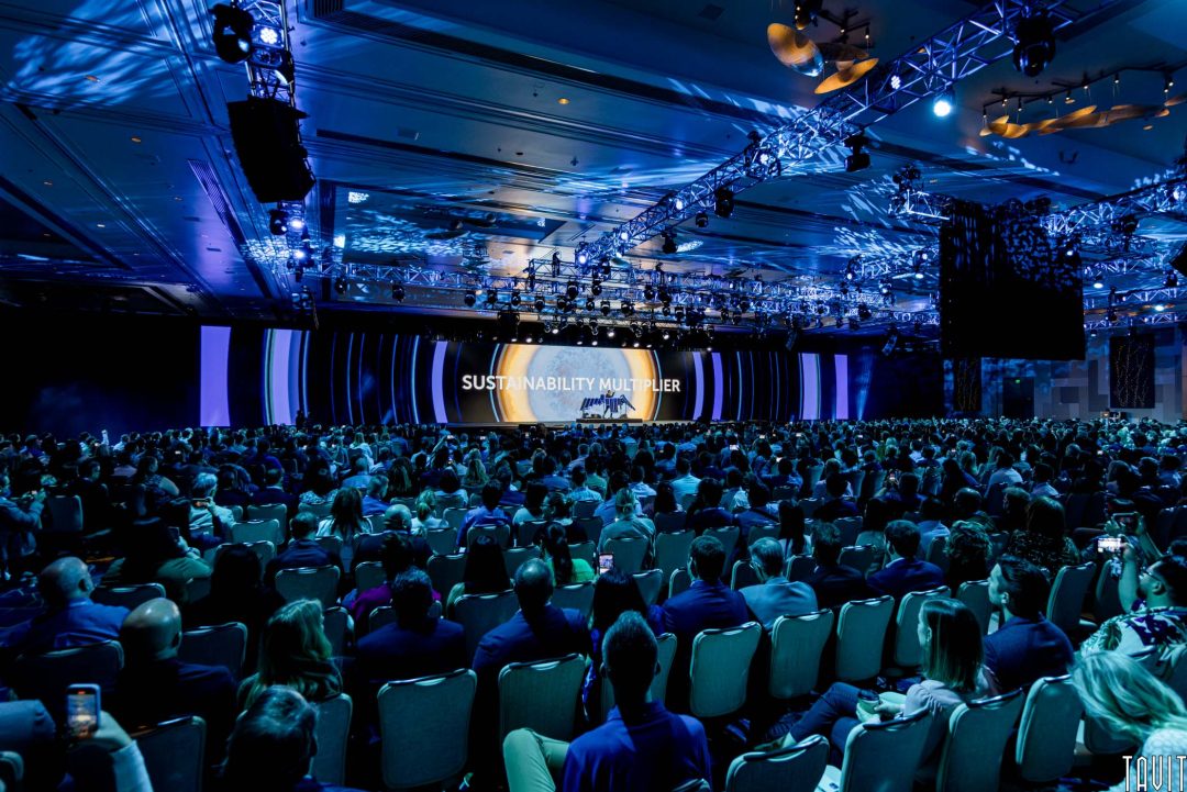 A large audience sits in a dimly lit conference hall at a corporate event, facing a stage with the words "SUSTAINABILITY MULTIPLIER" projected on a screen, surrounded by blue lighting and decorative ceiling lights.