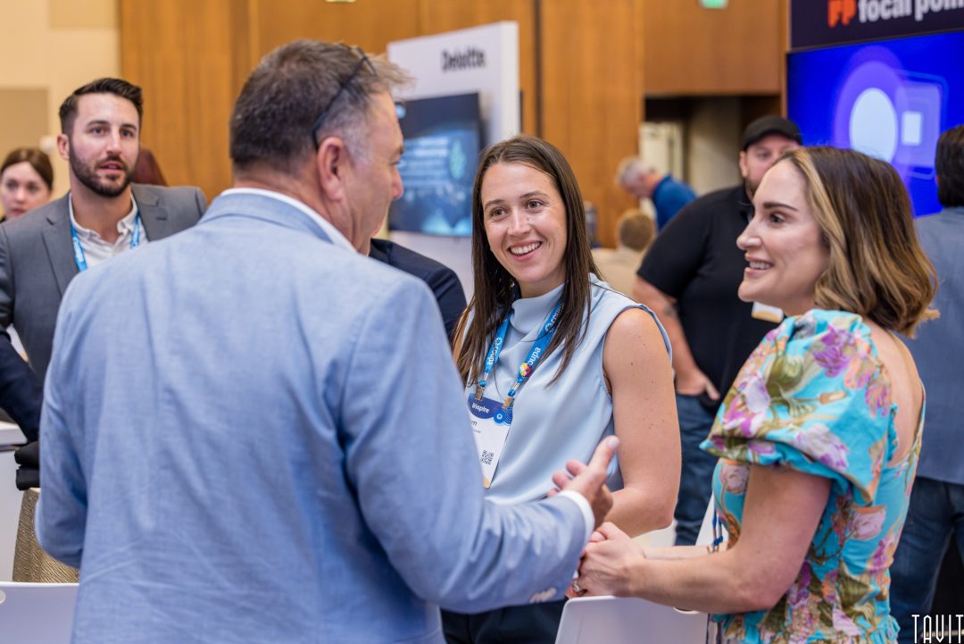 A group of people stands indoors at a corporate event, conversing and smiling. A man in a suit faces two women, one wearing a name badge and the other in a floral dress. Other attendees are visible in the background.