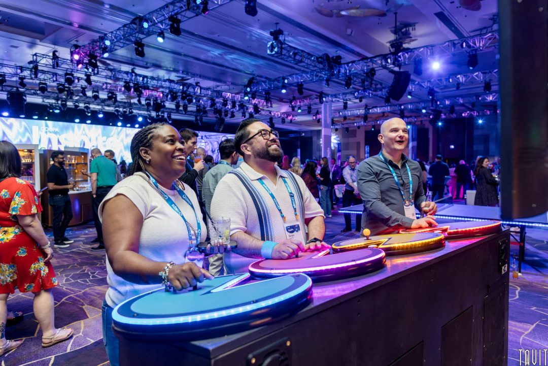 Three people happily play an arcade game with large colorful buttons at a lively corporate event, surrounded by other attendees and bright lights.