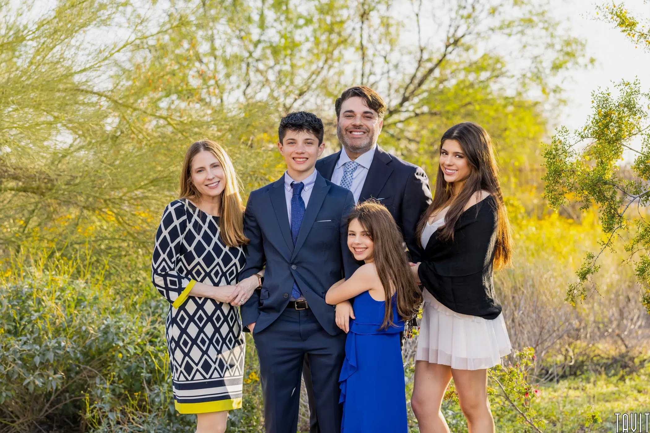 A family of five poses outdoors, smiling at a corporate event. There are two adult women, one adult man, a teenage boy in a suit, and a young girl in a blue dress. Greenery and sunlight fill the background.