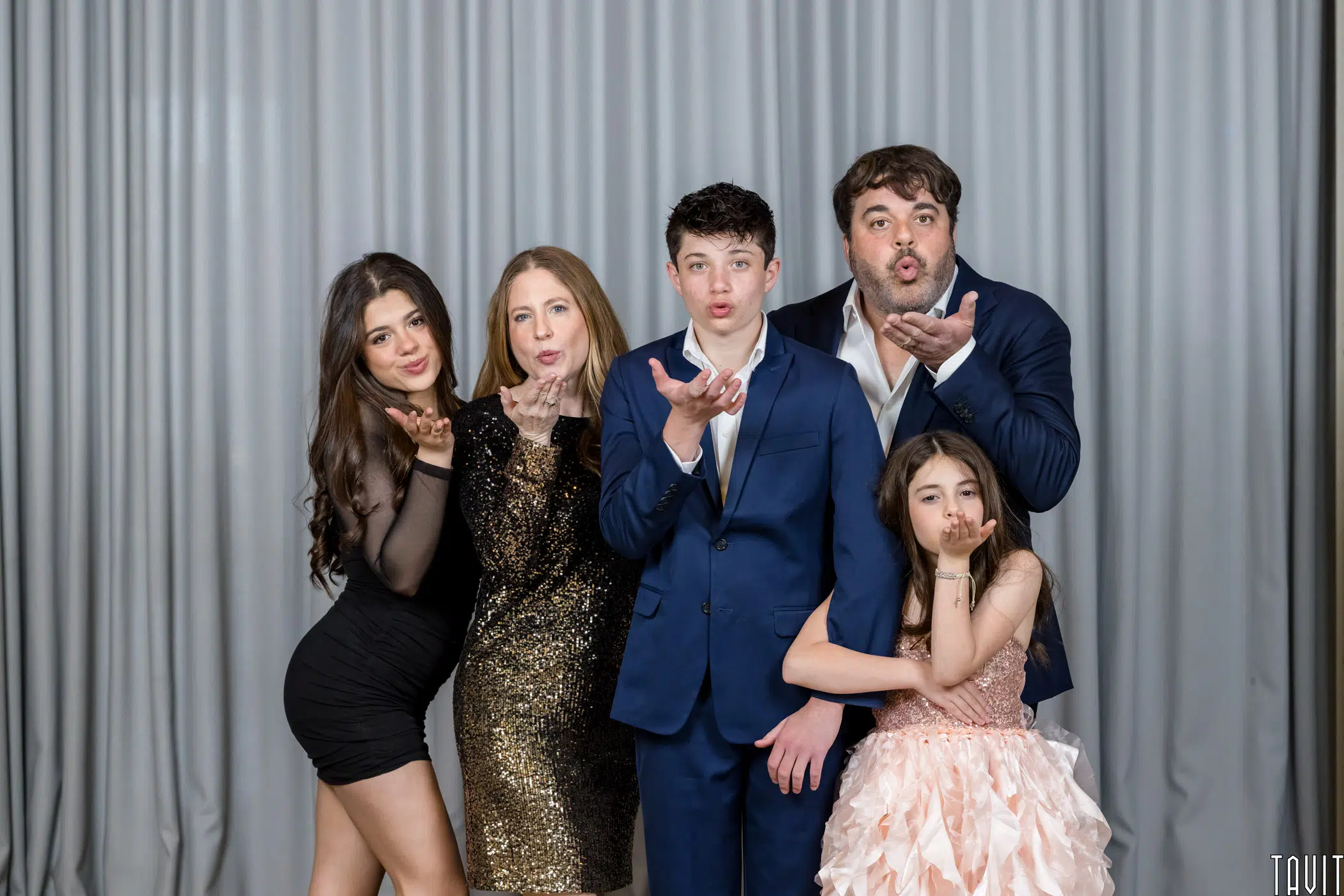 Five people dressed in formal attire stand in front of gray curtains, blowing kisses toward the camera at a corporate event. The group includes three adults and two children, all smiling and posing playfully.