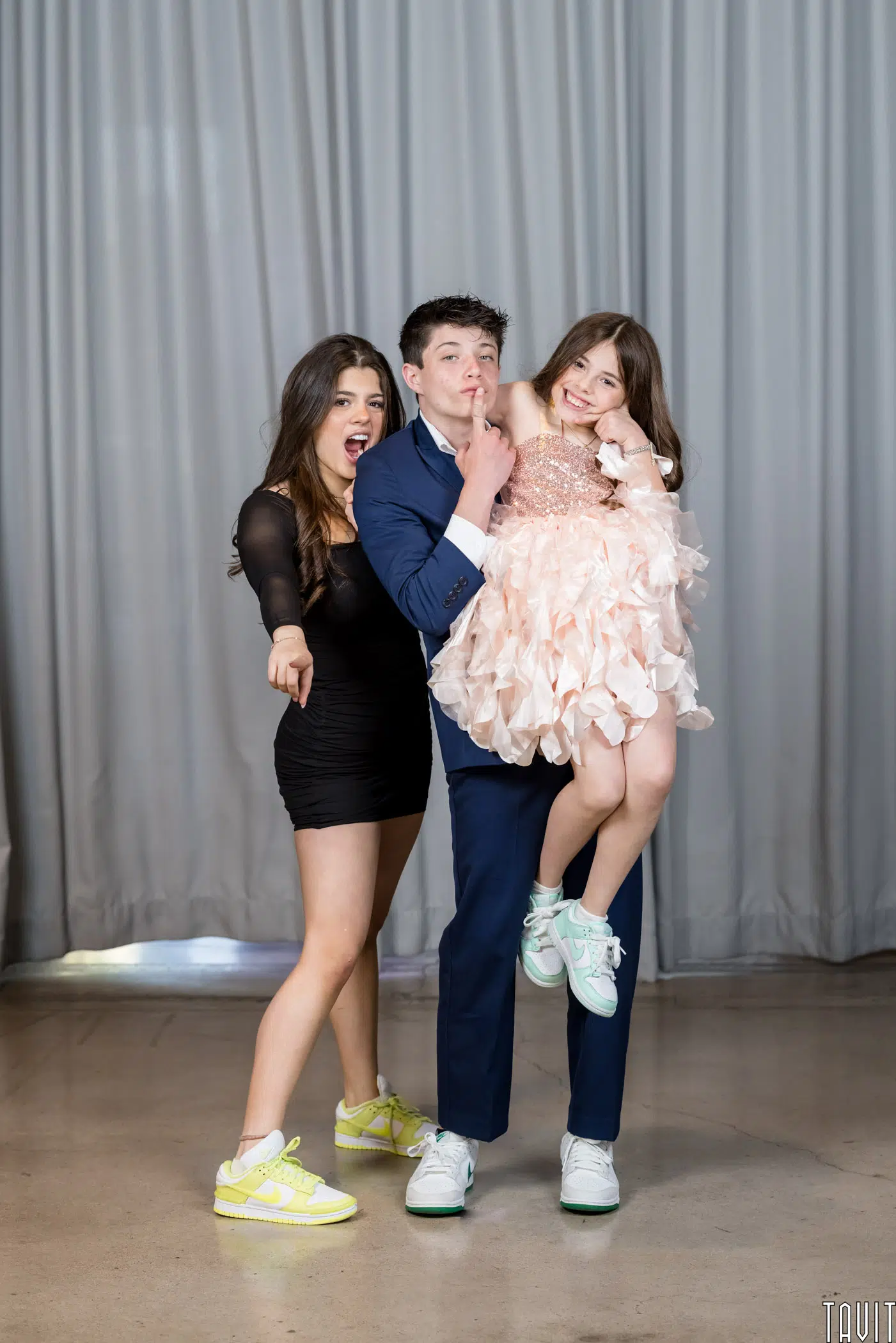 Three teens pose playfully in front of grey curtains at a seminar. A boy in a suit carries a smiling girl in a pink ruffled dress, while another girl in a black dress and yellow sneakers points at the camera.