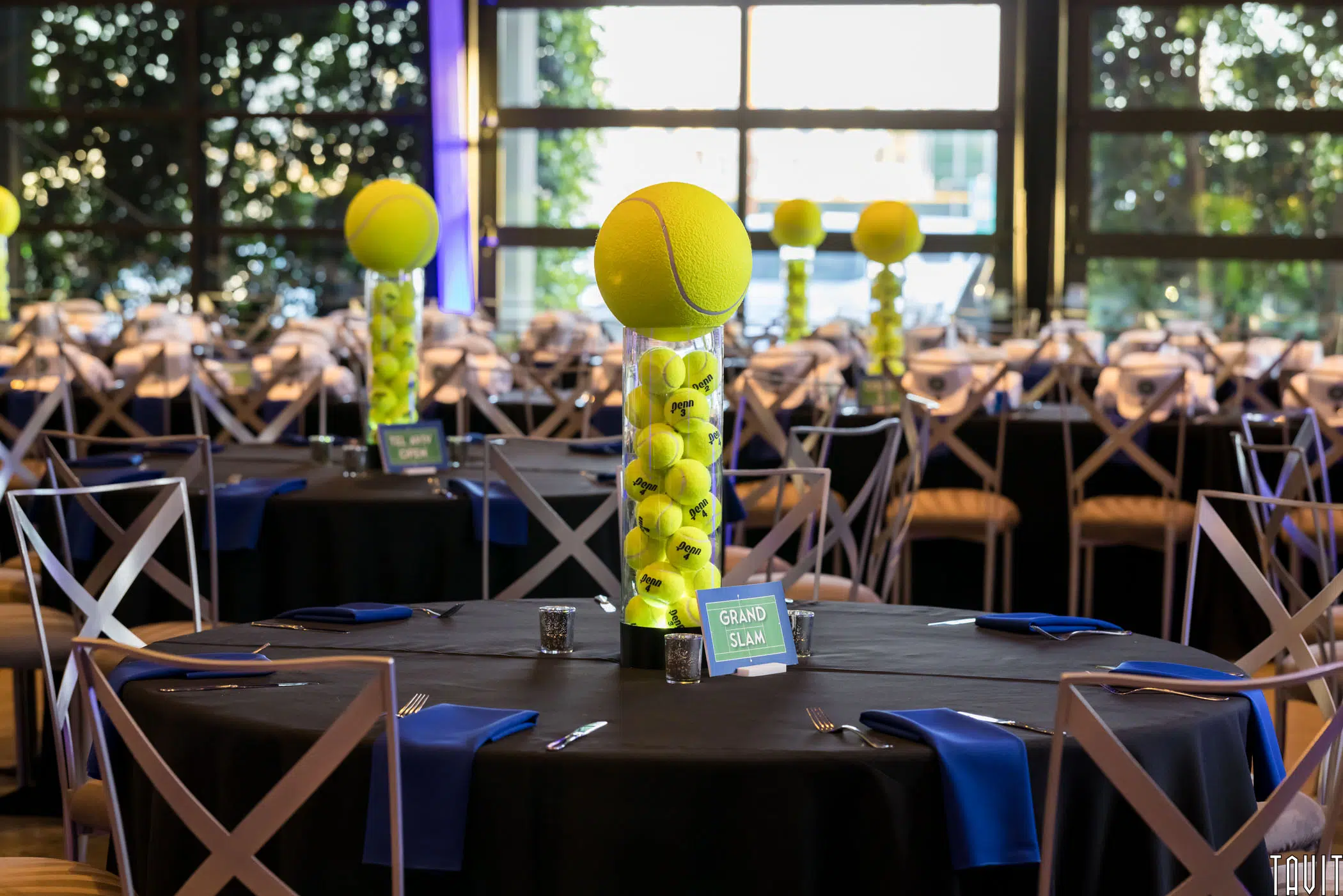 Round tables with black tablecloths and blue napkins are set for a corporate event. Each table features a striking centerpiece: stacked tennis balls in a tall glass vase, topped with a large tennis ball.