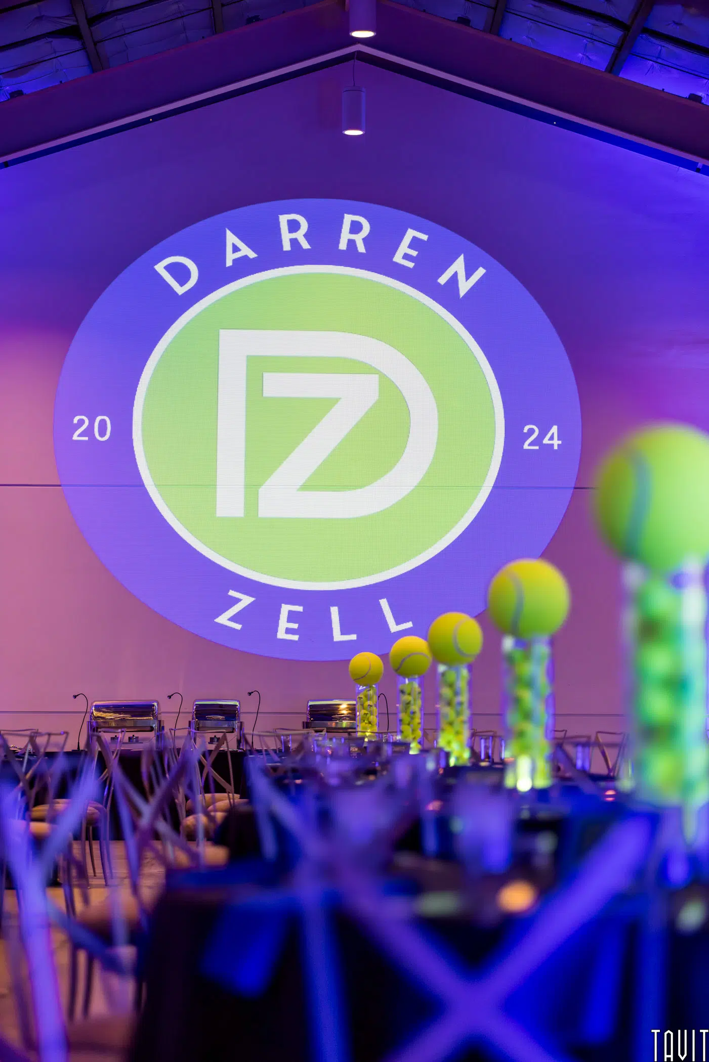 A modern event space perfect for a corporate event, decorated with tall centerpieces made from stacked tennis balls, and a large projected "Darren Zell 2024" logo glowing on a purple-lit wall in the background.