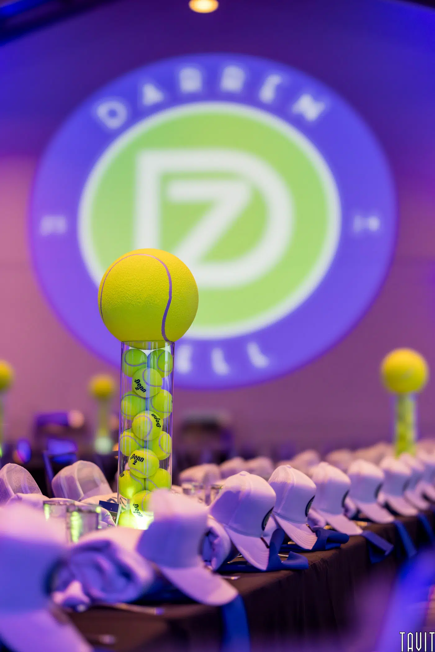A tall centerpiece with a large tennis ball and smaller tennis balls inside a clear cylinder sits on a banquet table lined with white caps at the corporate event. A circular neon "DARREN" logo is projected on the wall in the background.