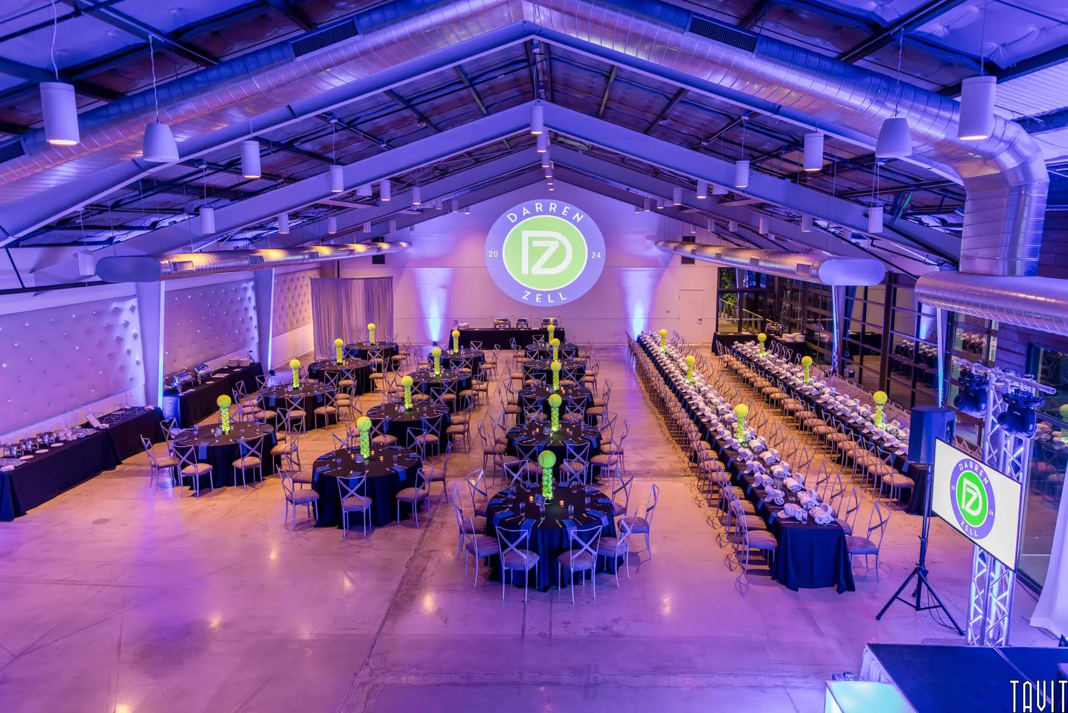 A modern event hall set up for a corporate event or seminar, with round and long tables decorated with green centerpieces and a large projection of a logo reading “DARREN ZELL” on the back wall under purple lighting.