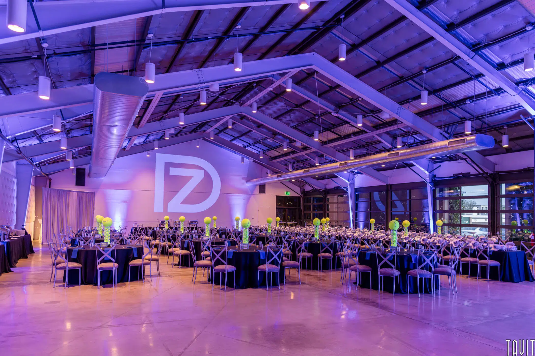 A spacious, modern event hall with high ceilings is set up for a corporate event or banquet, featuring round tables with black tablecloths, cross-back chairs, green balloon centerpieces, purple lighting, and a large "DZ" logo projected on the wall.