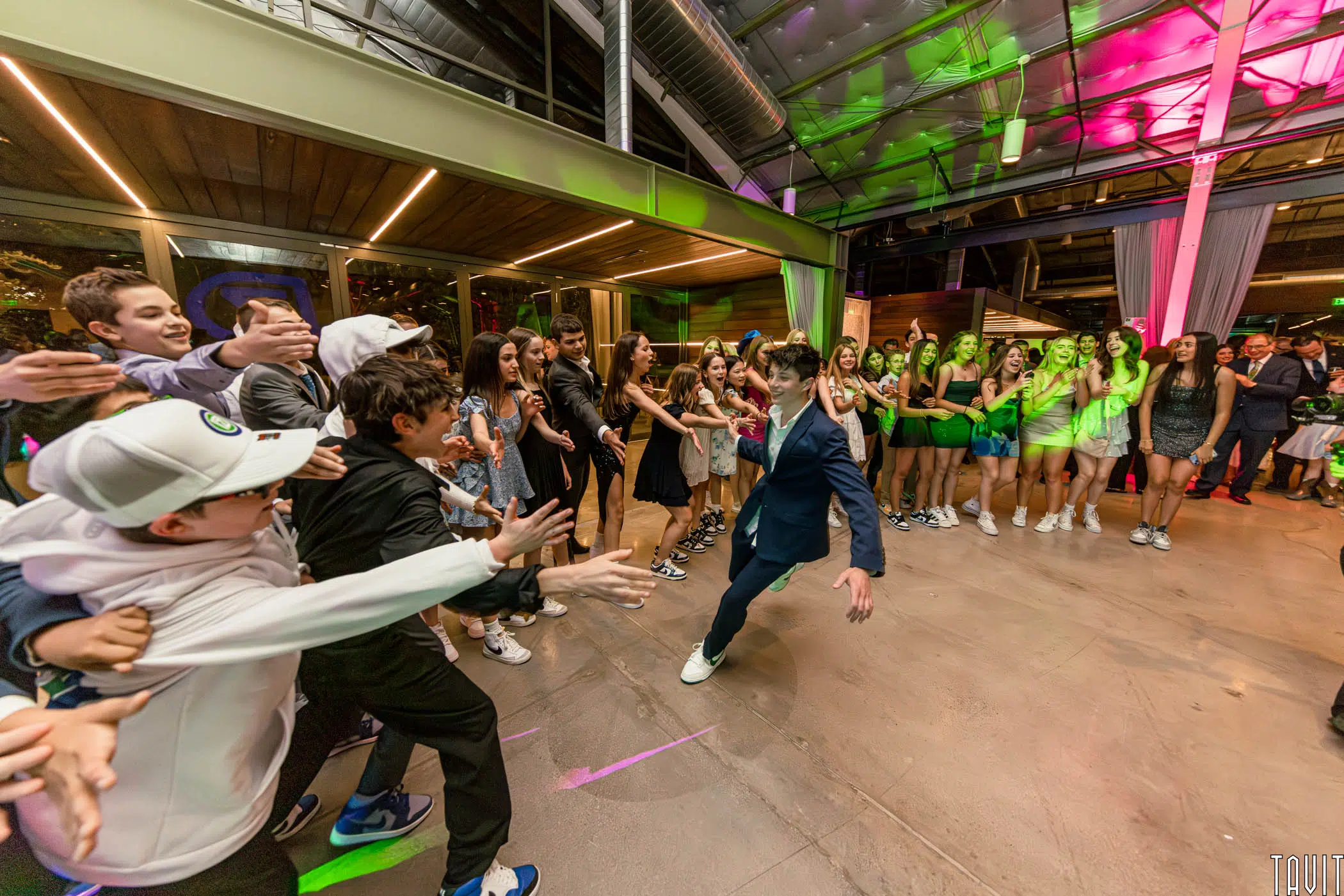 A boy in a blue suit dances energetically in the center of a corporate event, while excited friends in casual clothes reach out toward him. A crowd of teens watches, smiling, under colorful lights in a modern indoor venue.