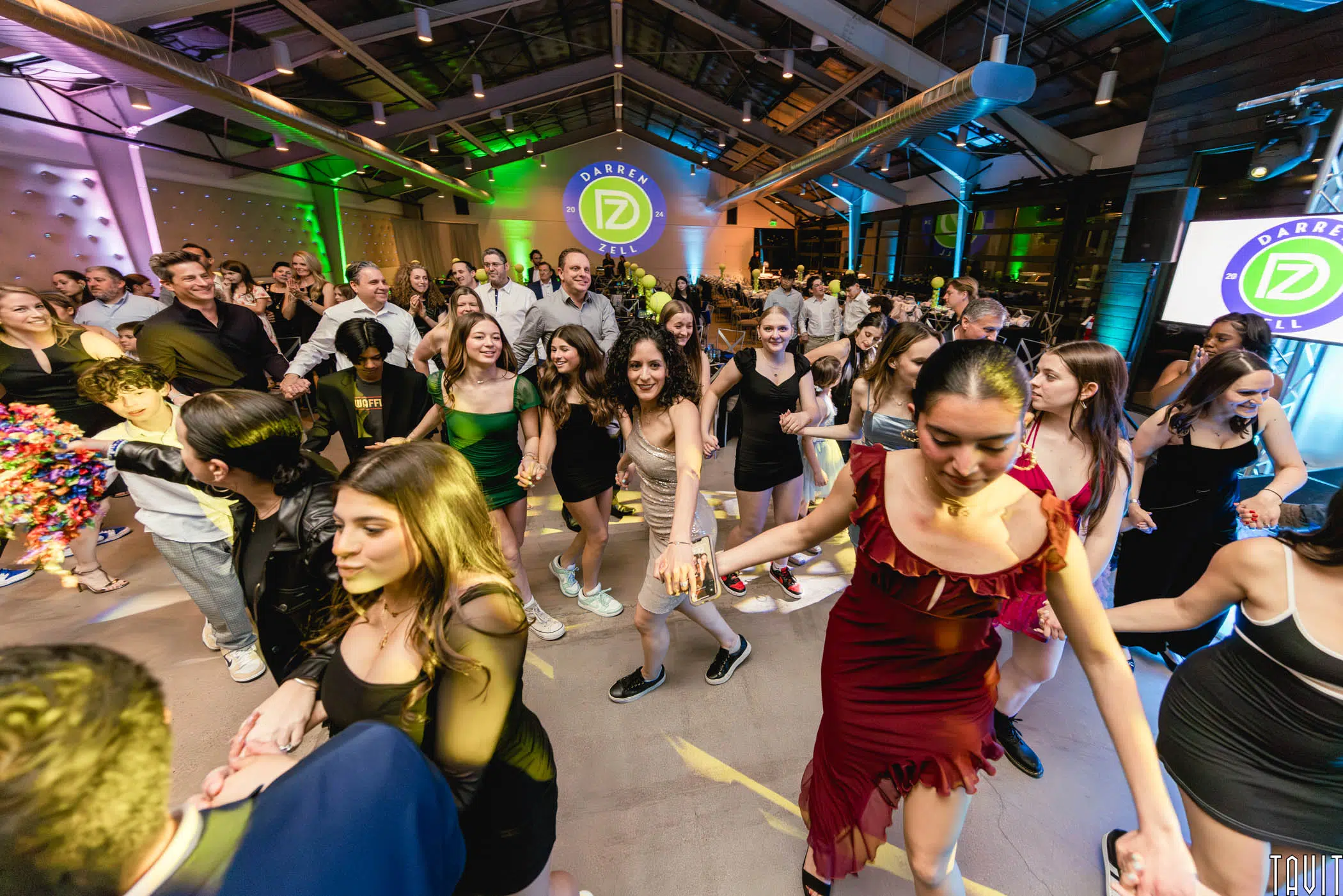 A large group of people, many smiling, dance together indoors under colorful lights at a modern corporate event. The venue features high ceilings, and event branding is visible on circular screens in the background.