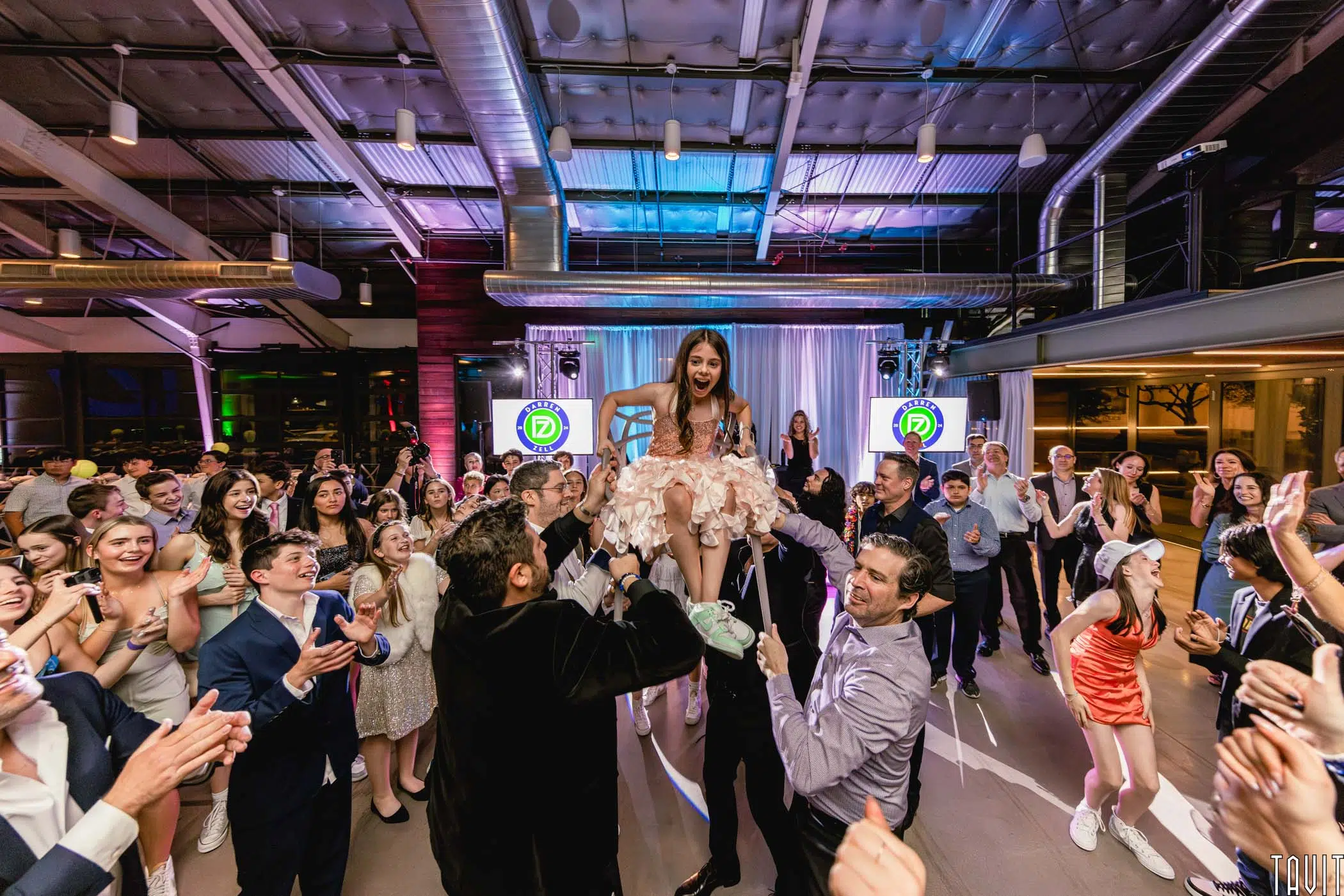 A young girl in a pink dress is lifted on a chair by several men while a large group of people surrounds her, clapping and celebrating at a lively expo venue decorated with lights.