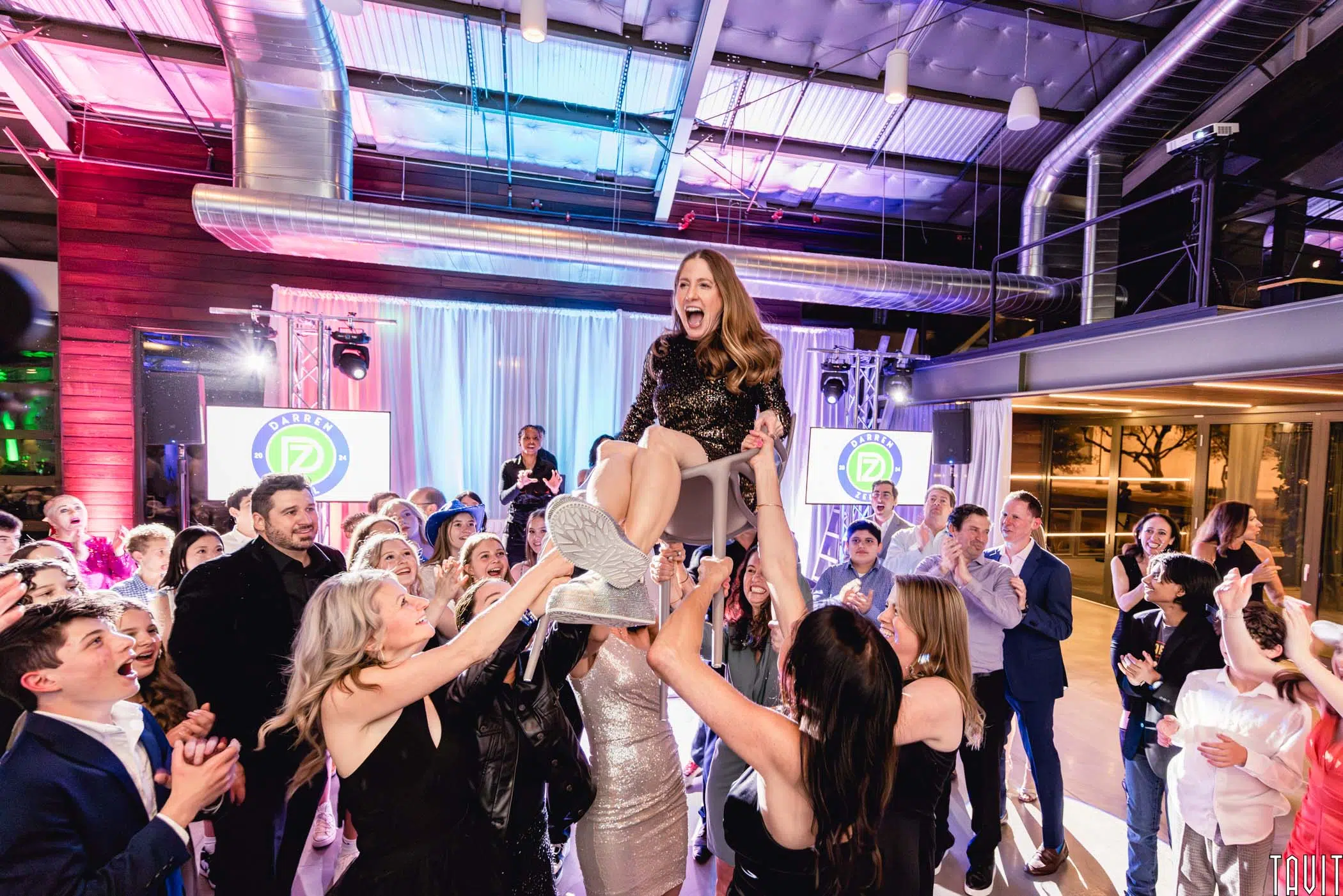A smiling woman is lifted on a chair by a group of people at a lively corporate event, with cheering guests and colorful lights in the background.
