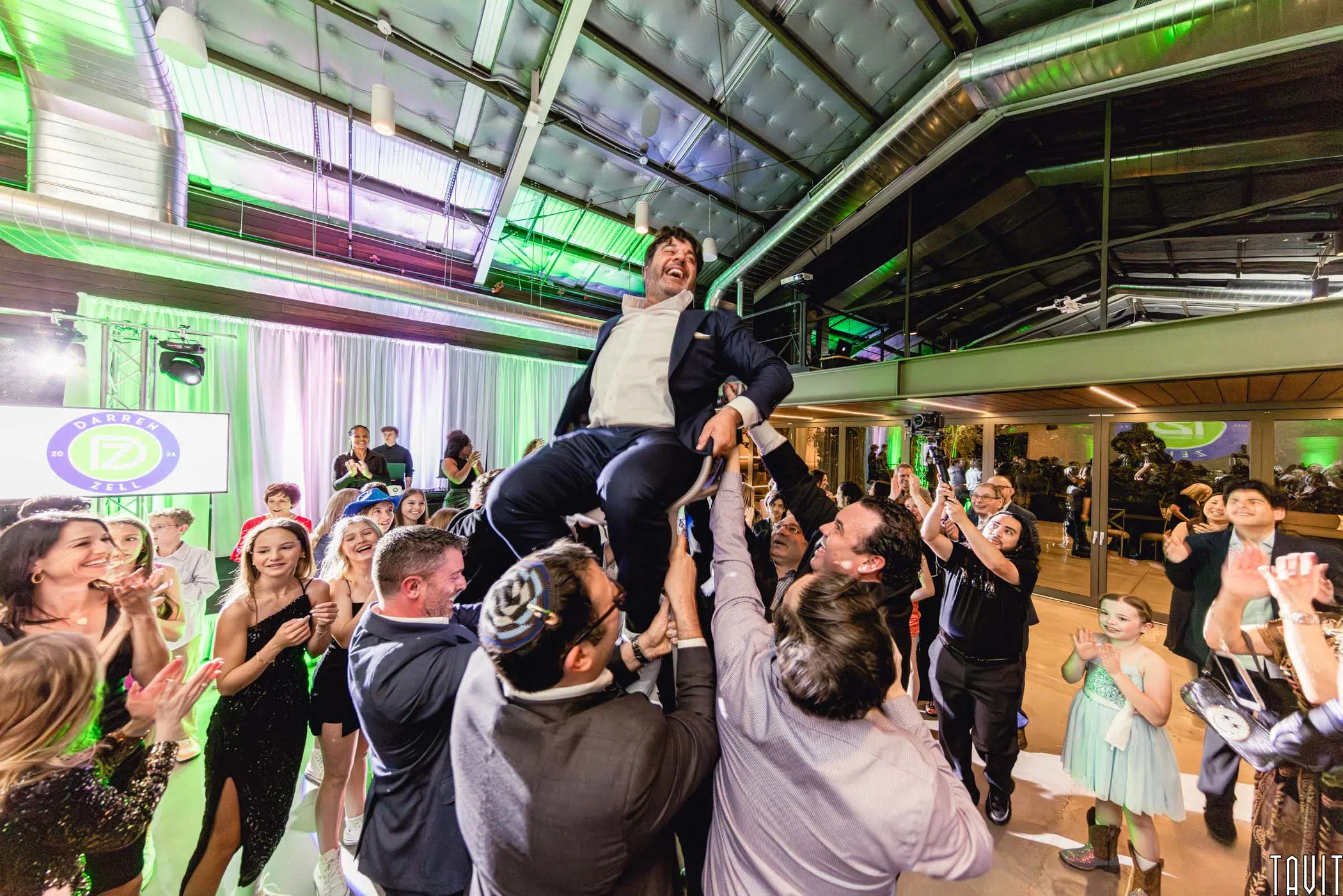 A group of people lift a smiling man in a suit on a chair during a lively corporate event indoors, while guests cheer, clap, and dance around them under bright lights.