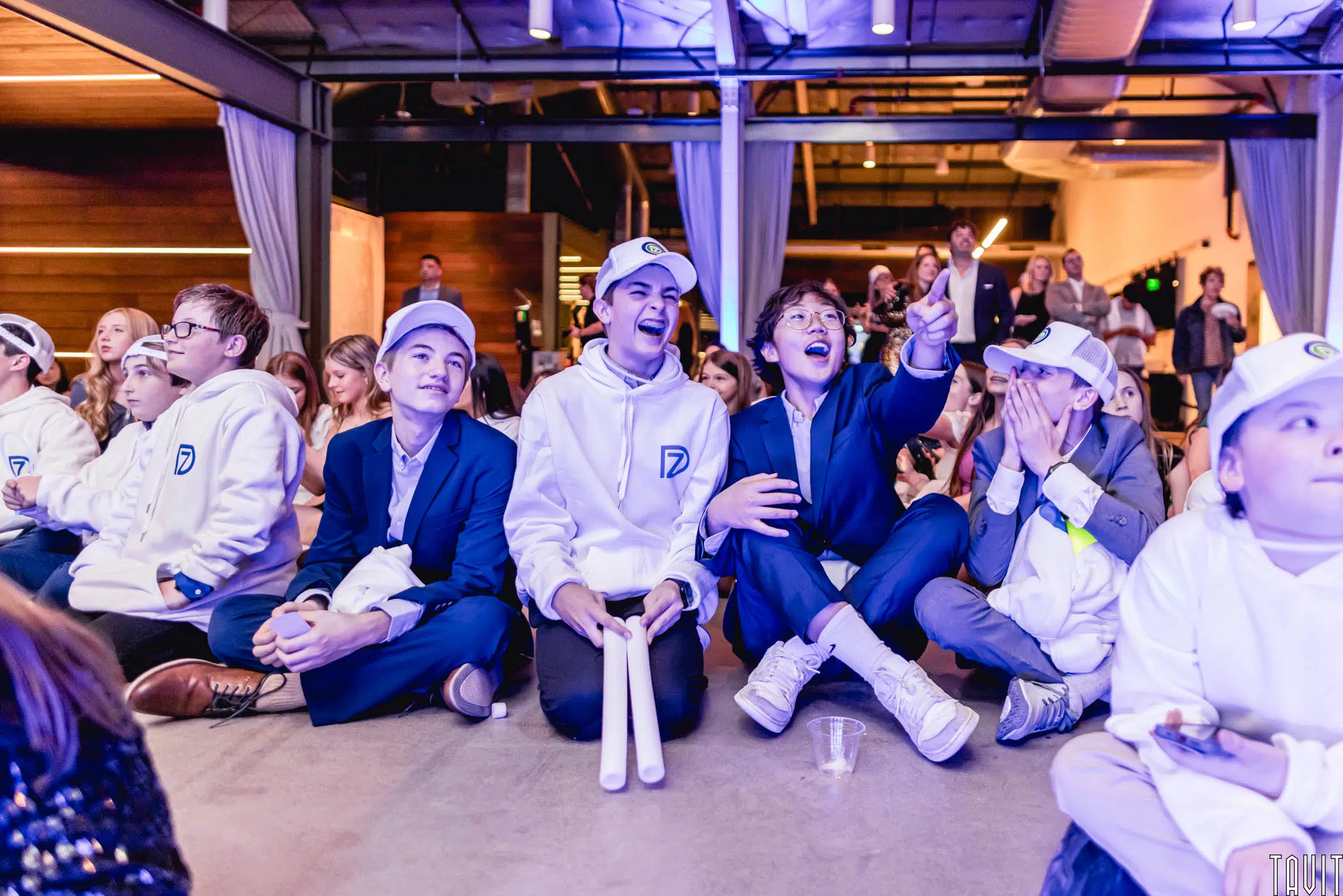 A group of excited children and teens, some in white hoodies and caps, sit on the floor at an indoor seminar, cheering and smiling under bright lights.