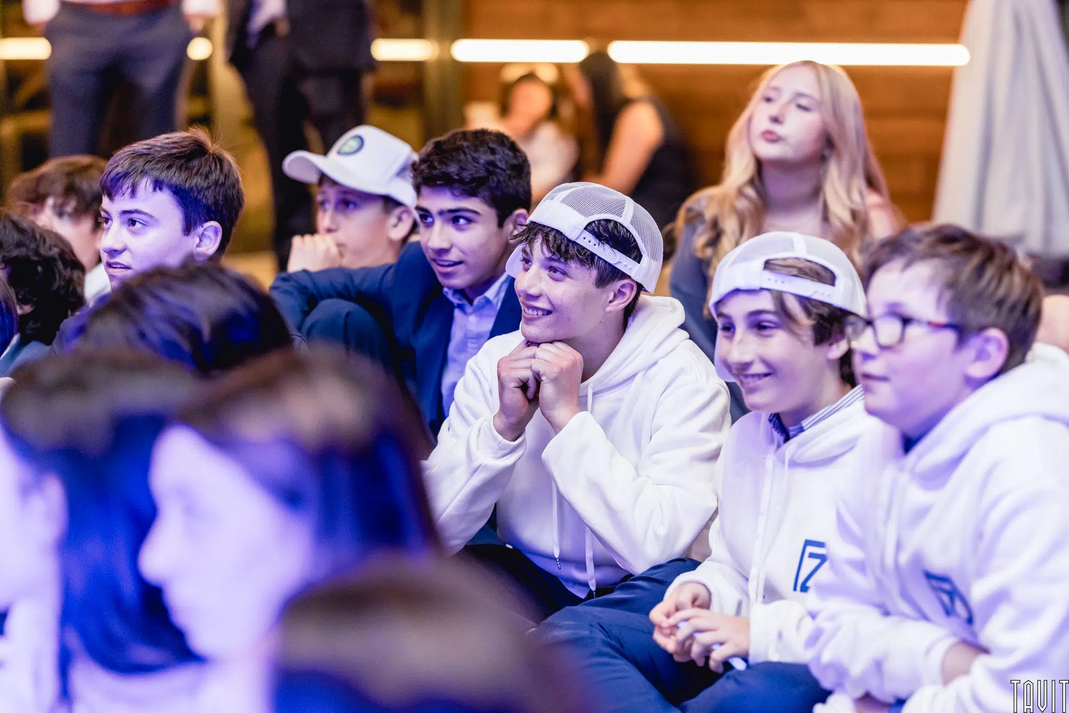 A group of boys wearing white hoodies and caps sit closely together, smiling and looking ahead during a seminar, while others sit around them. The background is softly lit, with people slightly out of focus.