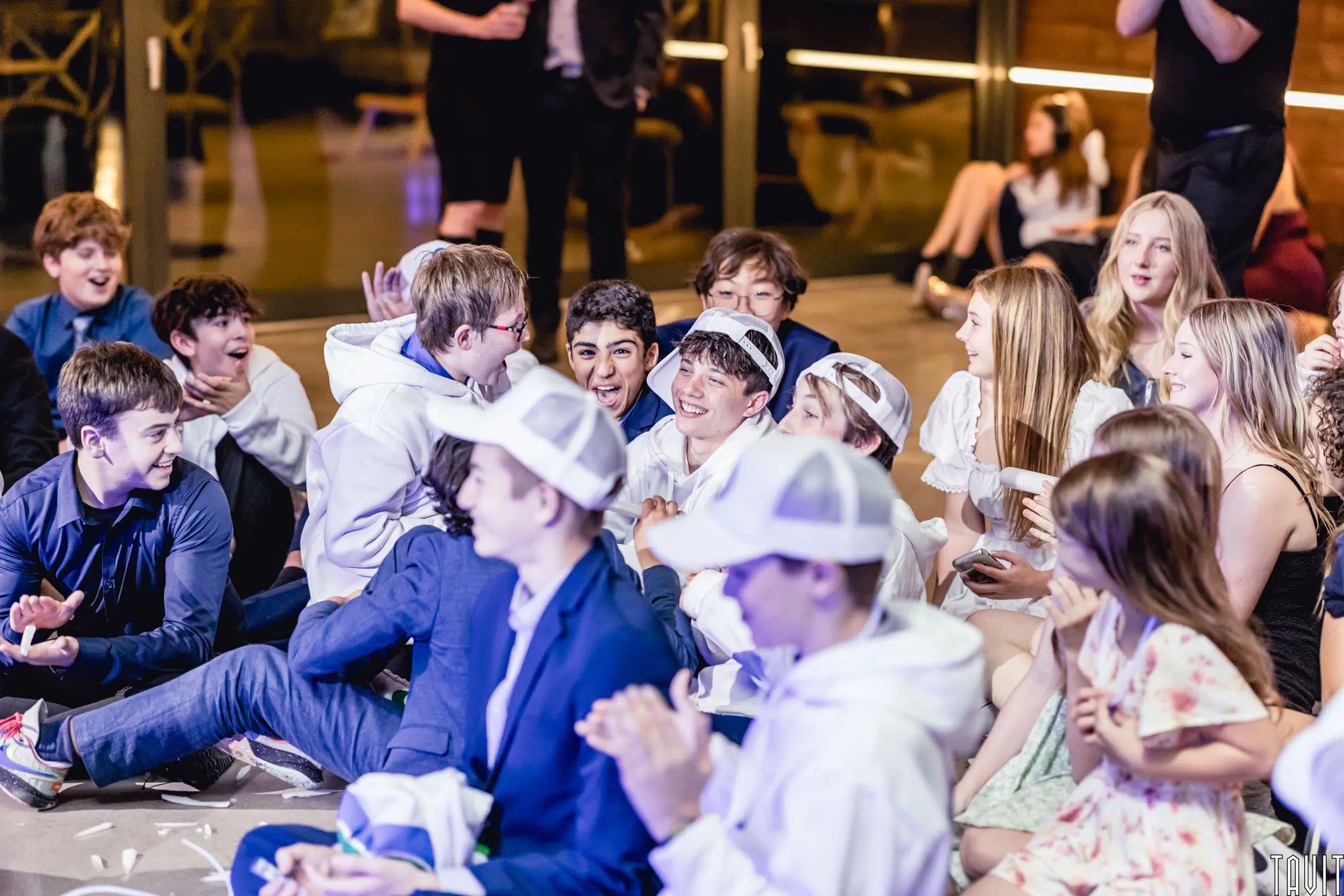 A group of teenagers, some in white hoodies and caps, others in suits or casual clothes, sit closely together on the floor at a lively seminar, smiling, laughing, and clapping.