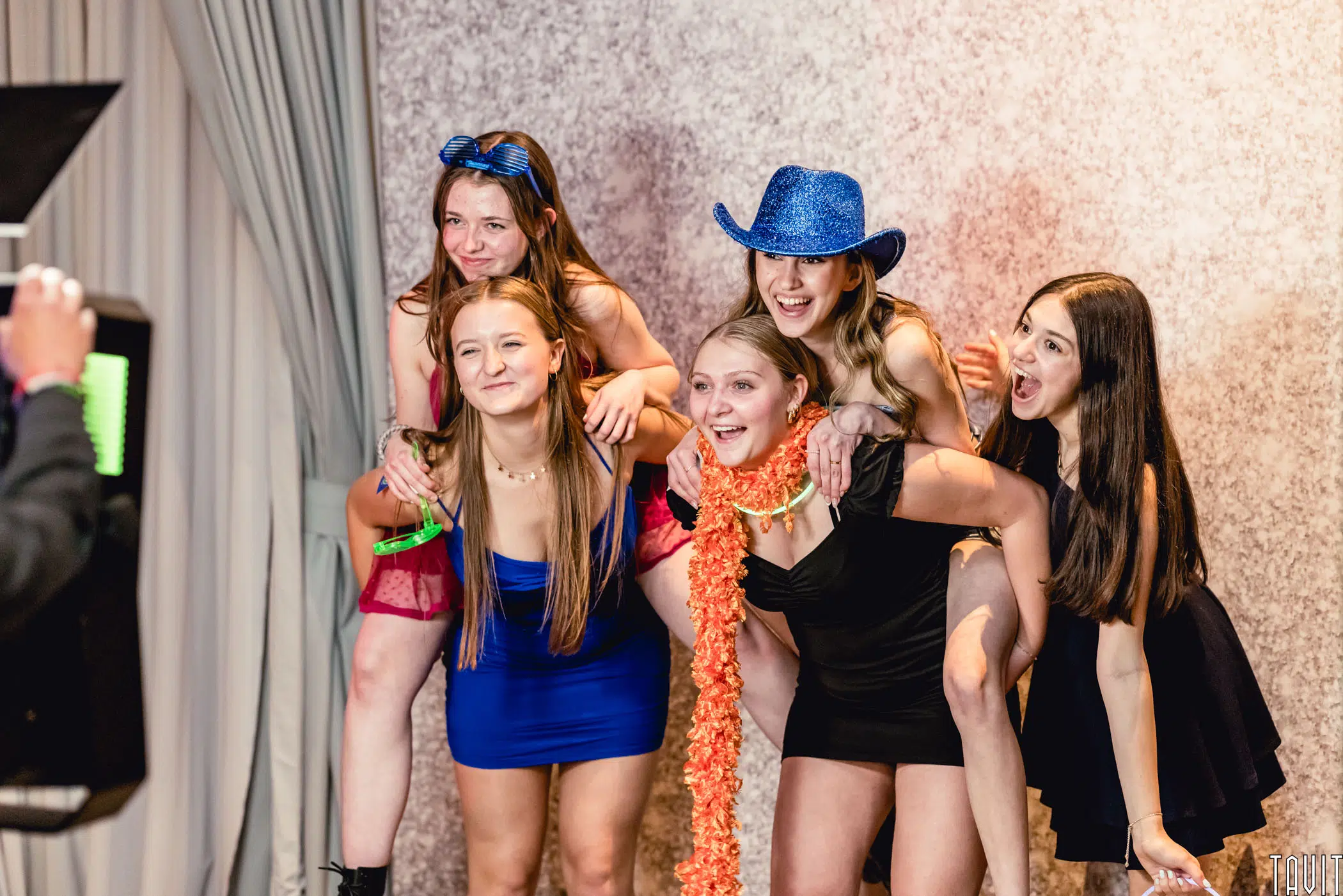 Five young women pose playfully together at a corporate event in front of a textured backdrop. Two crouch in front while three climb on their backs, all smiling and dressed for a party—one sporting a blue cowboy hat, another an orange feather boa.