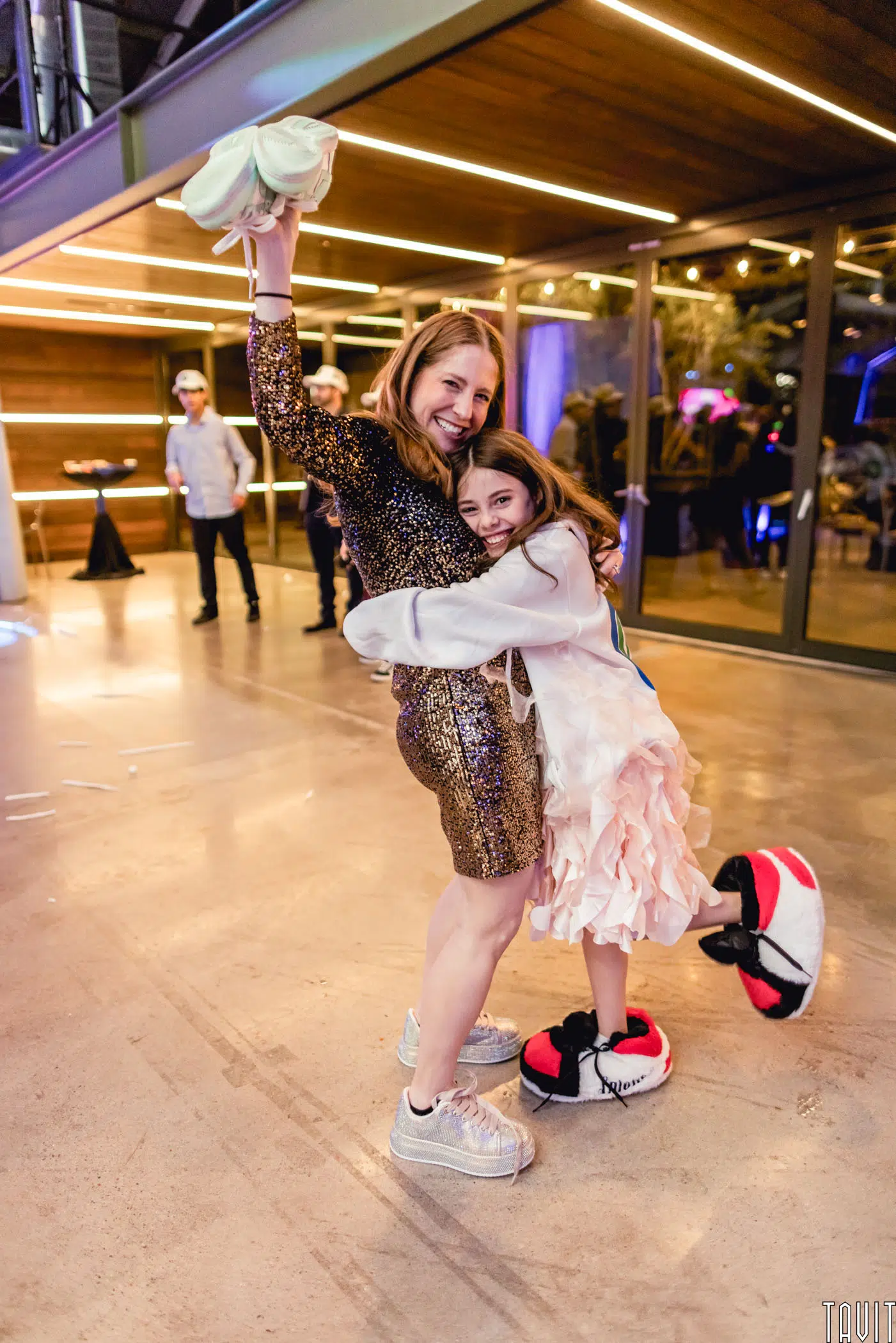 A smiling woman in a sparkly dress hugs a laughing girl in a ruffled dress. Both wear oversized novelty slippers and look joyful at an indoor corporate event with glass walls and string lights.