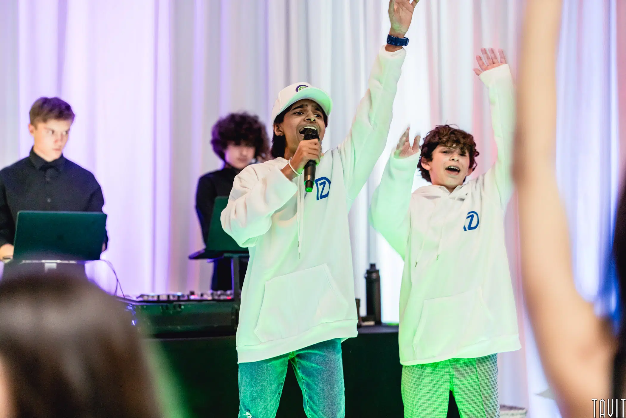 Two kids in white hoodies sing and raise their hands on stage, while two others stand behind DJ equipment. The background features white drapes and colorful lighting, creating a lively atmosphere perfect for a corporate event or expo.