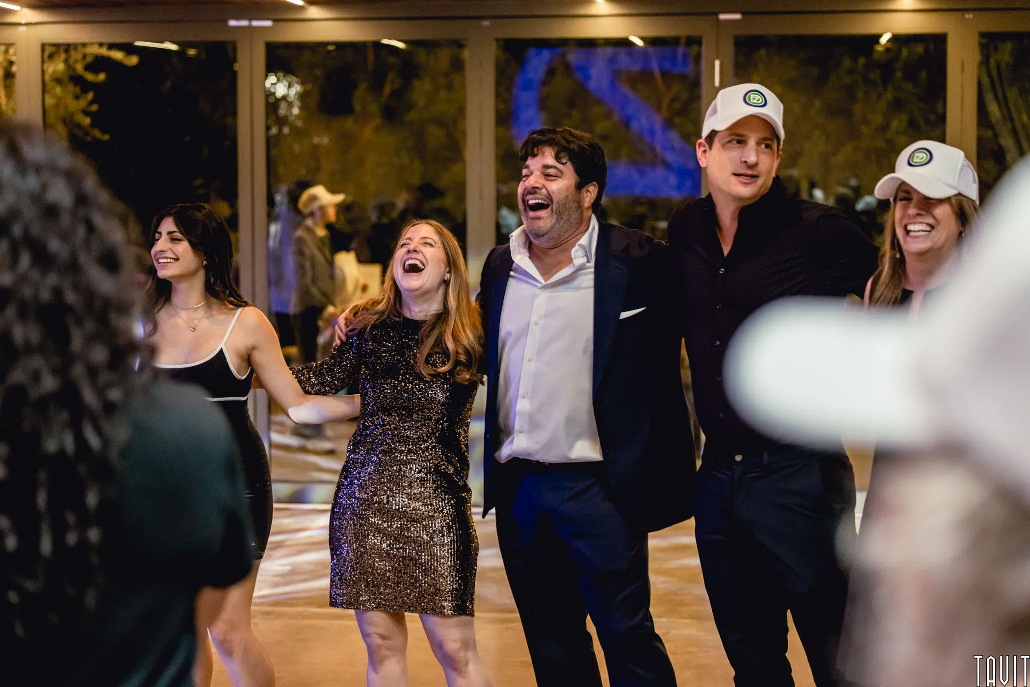 A group of four people stand side by side, smiling and laughing with their arms around each other at an indoor corporate event. Two wear formal outfits; two wear baseball caps. Other guests mingle in the background.