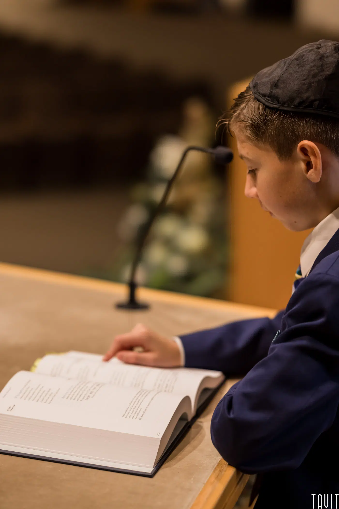 A boy wearing a kippah and suit reads from an open book at a podium, speaking into a microphone as if presenting at a seminar, with a blurred background behind him.