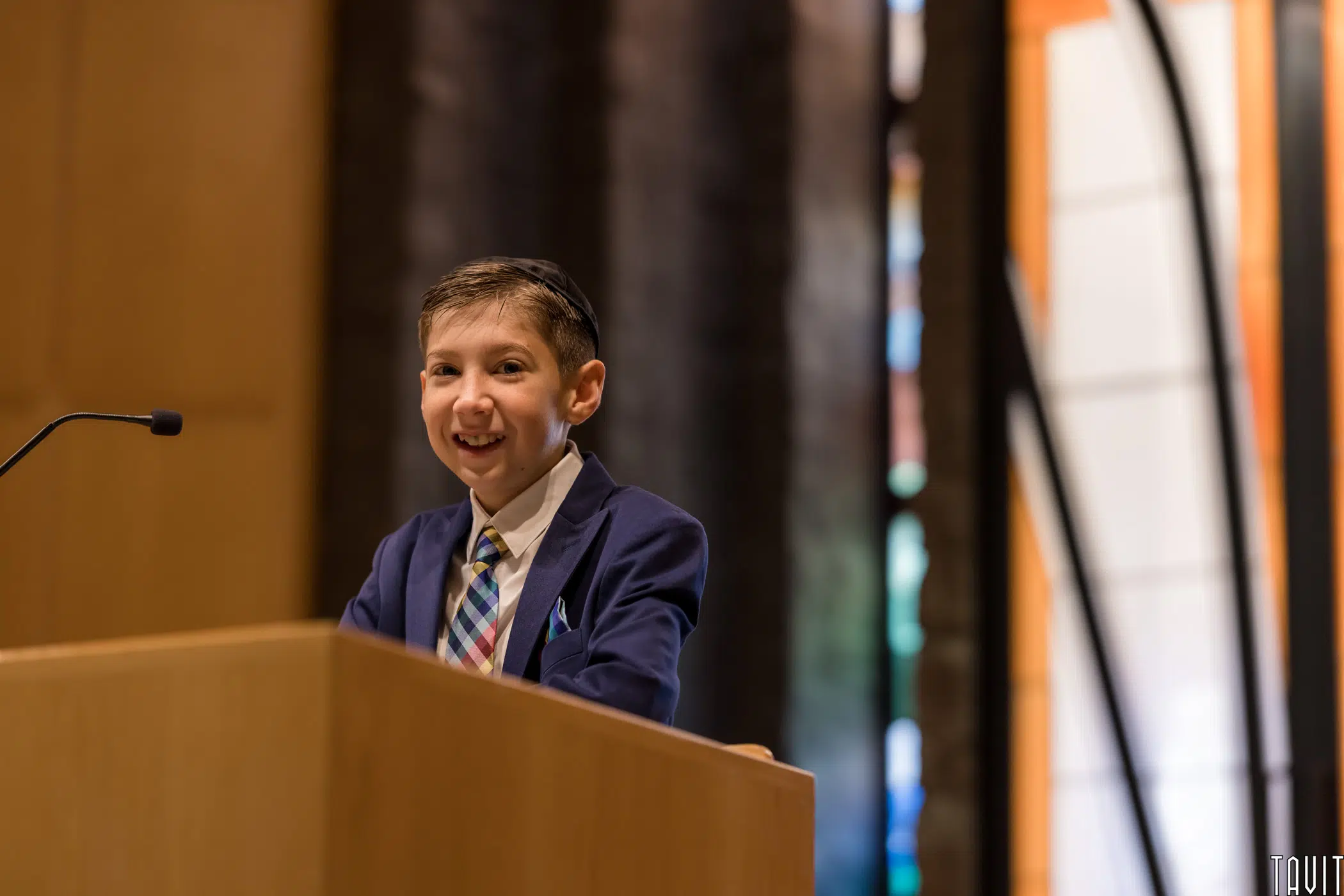 A young boy wearing a suit and tie smiles while standing at a podium during a seminar, with a microphone in front of him and colorful stained glass windows in the background.
