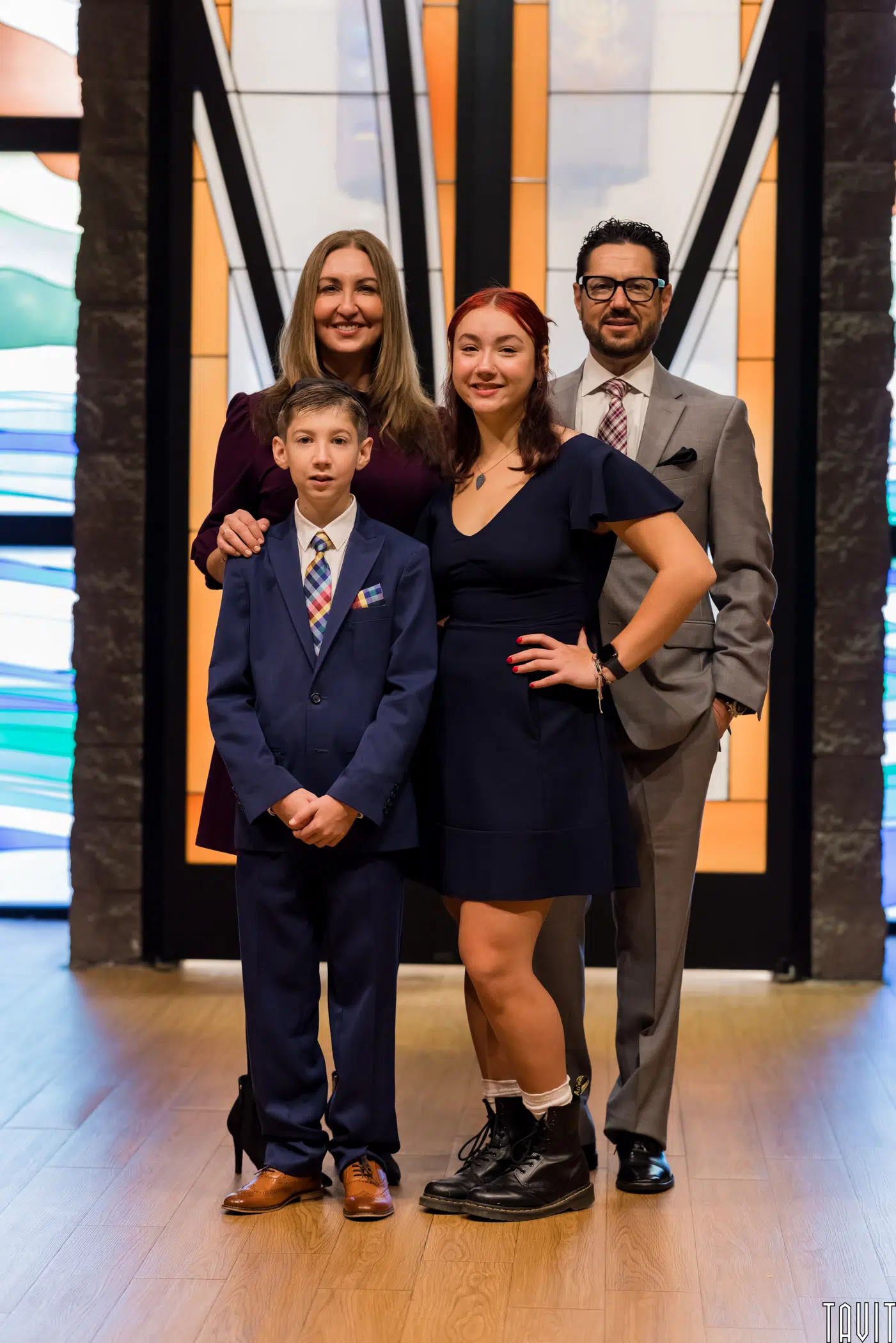A family of four poses indoors, smiling, at a corporate event. An adult woman and man stand behind a teenage girl in a dark dress and boots and a young boy in a suit. Stained glass windows are visible in the background.