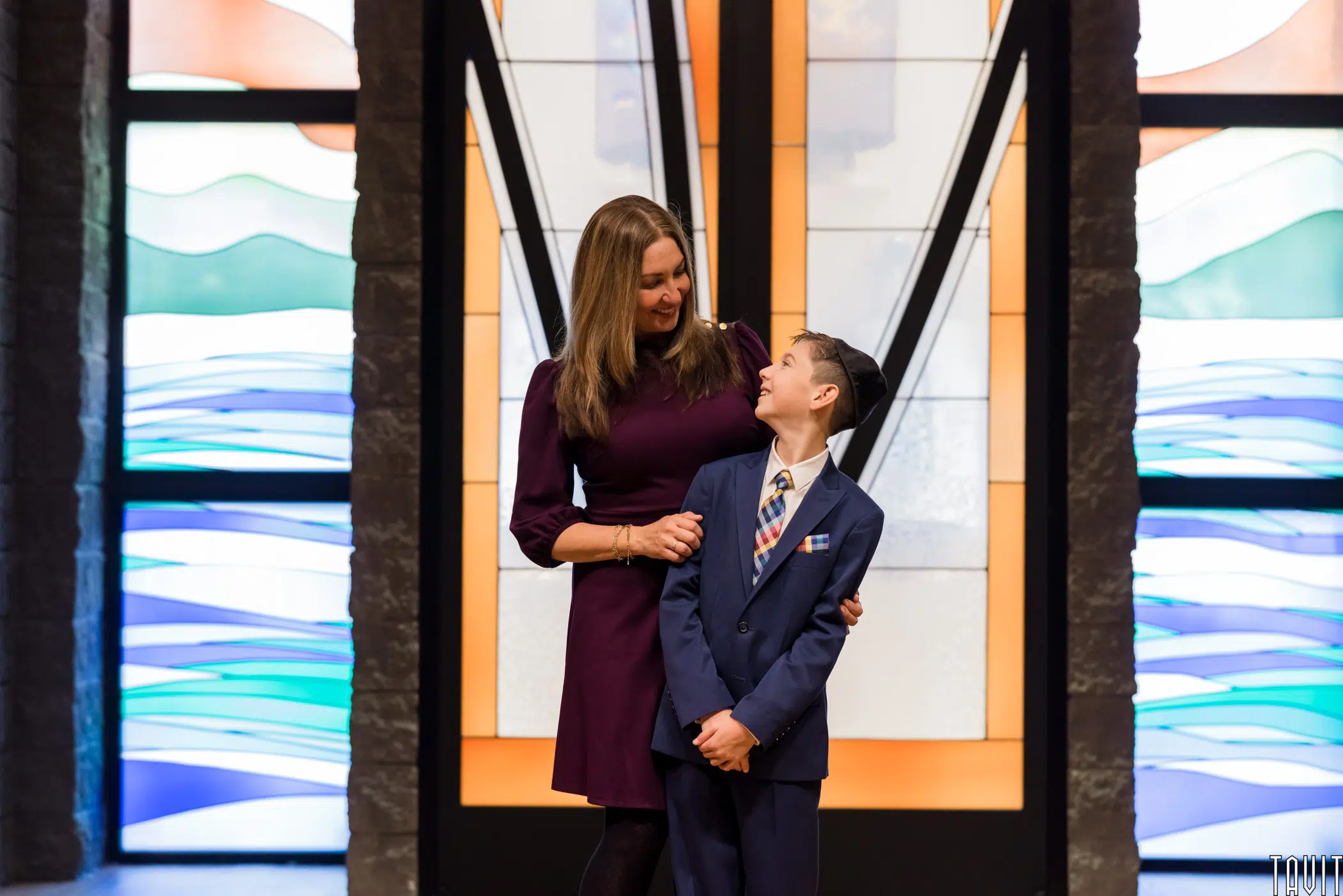 A woman in a purple dress stands with her arm around a young boy in a blue suit and multicolored tie. They are smiling at each other at a corporate event, with vibrant stained glass windows behind them.